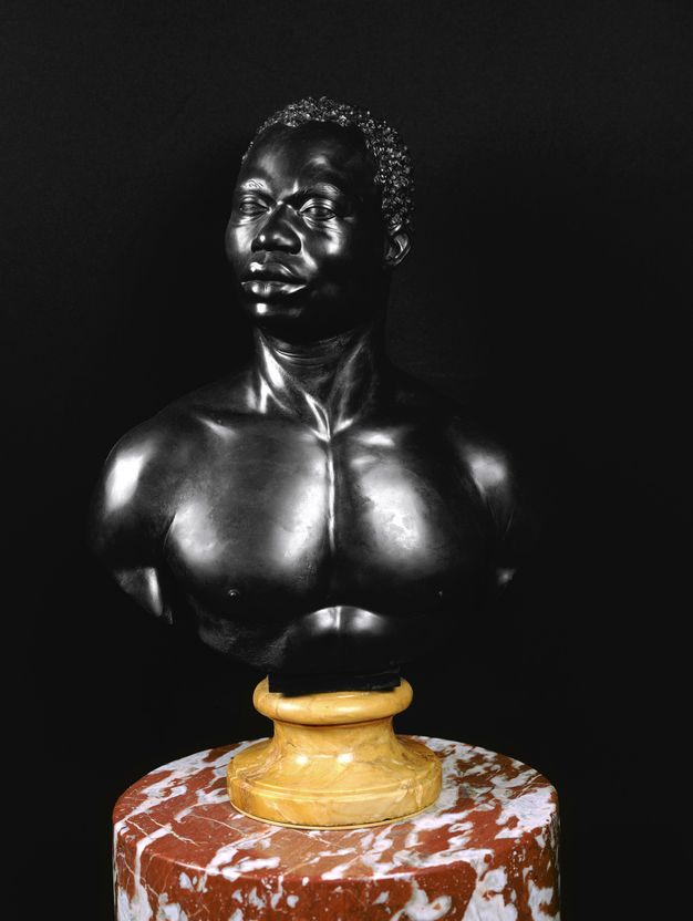Photograph. Sculpted bust shows a glossy surface that contrasts sharply with the ink-black background. The yellow socle almost glows, and it sits on a red and white stone column.