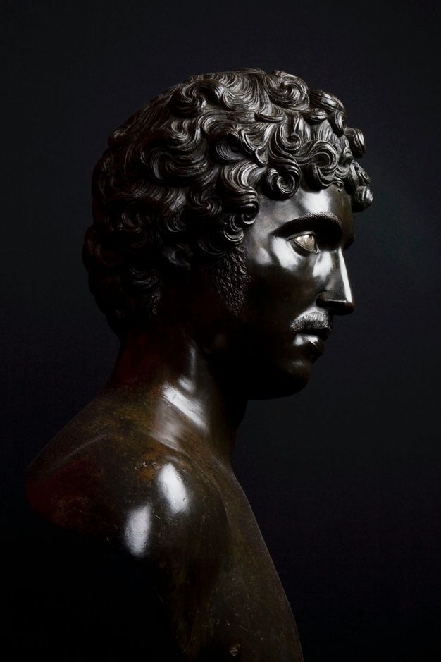 Photograph. Sculpted bust faces our right in profile against a deeply shadowed background. Light gleams off the polished bronze surface.