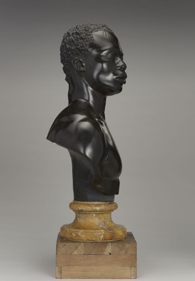 Photograph. A sculpture faces our right in profile. The back cuts in where the shoulder blades would be to meet the yellow socle. The whole bust sits up on a wooden block in this photograph.
