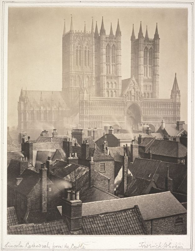 Black and white photograph. In tones of warm brown and grey, the pointed towers and spires of a cathedral rise high above the nearby rooftops. Across the town from us, the church is faint, almost otherworldly, in the distance.