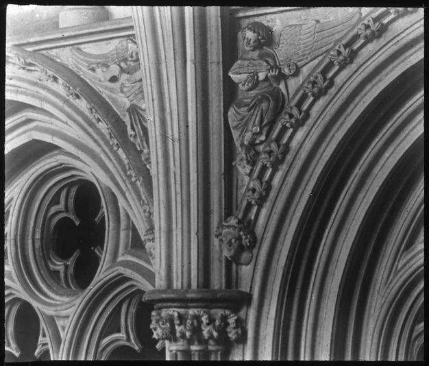 Black and white photograph. Close up view of the triangular area between a curving arch and a rib leading to the cathedral ceiling. Carved in relief there, a winged angel appears to close their eyes while strumming an instrument.