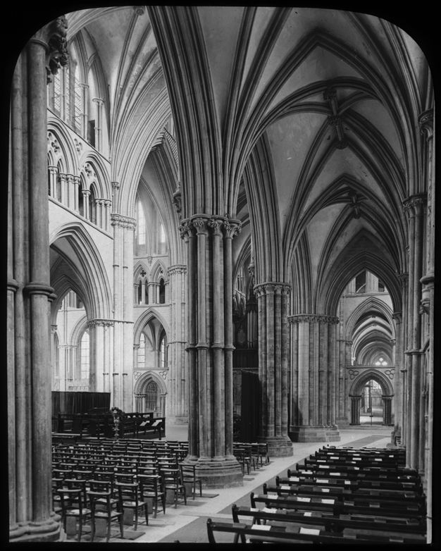 Black and white photograph. Held aloft by clustered columns, the groin vaults of a cathedral soar over chairs that seem diminutive by comparison.