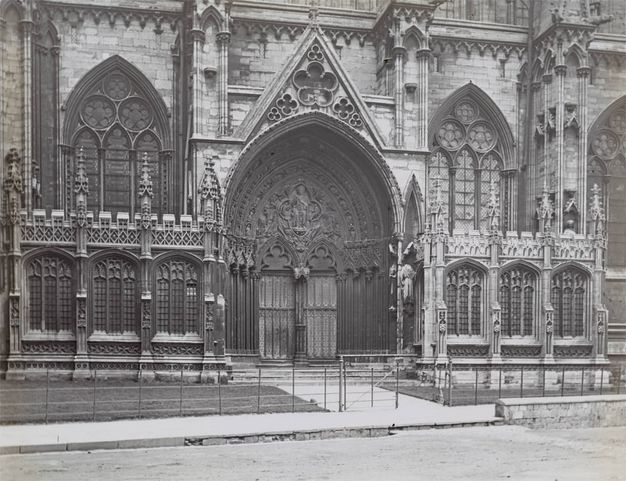 Black and white photograph. A double door on one porch of a cathedral is engulfed within nested arches, which are then crowned by a pointed pediment. Windows, buttresses, and rows of spiky pinnacles are heavily ornamented.