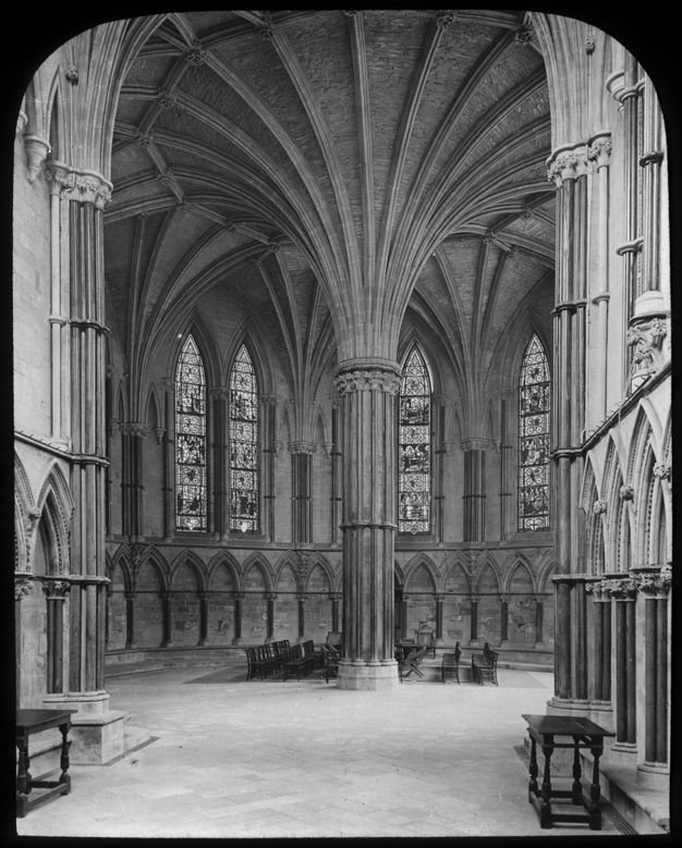 Black and white photograph. Flaring like a trumpet’s horn, ribs curve up and out of a central pier at the heart of a polygonal chapter house. The wall is lined with pointed, arched windows filled with stained glass.