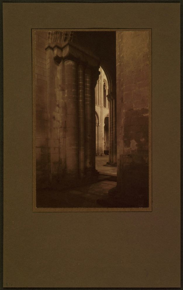 Sepia toned photograph. In tones of parchment and tawny brown, we look through a sliver of an opening into a light-filled nave of a cathedral. There are columns and rounded arches in both spaces.