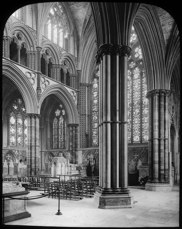 Black and white photograph. A retrochoir is backed by a stained-glass window at least several stories tall. Clustered columns support high, pointed arches, which run under the gallery level. Light pours in through clerestory windows and others covering nearly every other surface above and around the retrochoir.