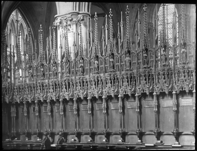 Black and white photograph. Individual seats along a bench-like row of a choir stall are separated by arms that project from a high back. Using the chairs for scale, the screen appears to be one or two stories high, and it is elaborately carved with tracery, spires, and figures.