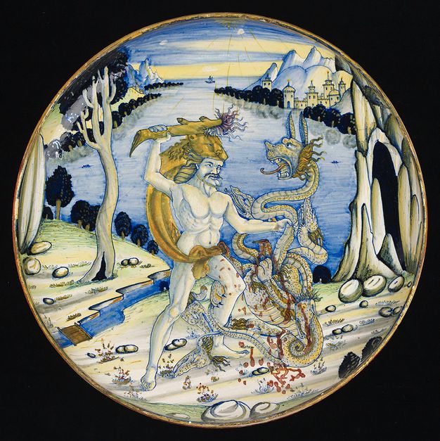 Dish. In tones of vivid blue, ochre brown, and rust red, a muscular Hercules raises a club to strike the serpent-like, multi-headed Hydra. Red blood pours from an already severed head. Hercules wears a lion’s pelt.