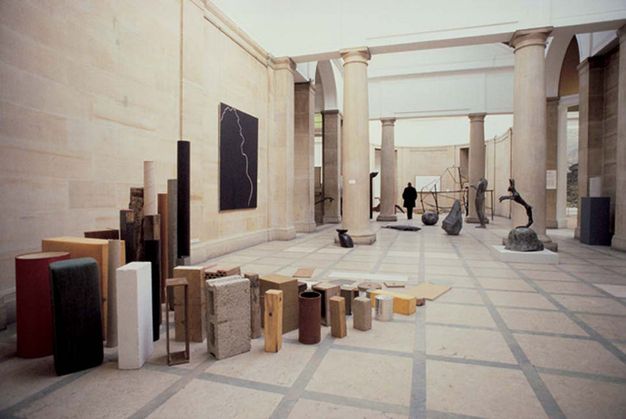 Installation photograph. Sculptures are placed around and across a stone floor in a columned room. The work closest to us, to our left, is made of what appears to be building and packing materials. There is one of each object, like one cinder block, one piece of lumber, one cardboard packing tube, one slab of marble, and one metal tube among other objects. They are arranged in descending size so loosely resemble breaking surf.