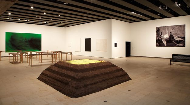 Installation photograph. Large-scale sculptures and a bench are spaced around a gallery, and the walls are hung with works of art. The piece closest to us is like a miniature stepped pyramid with brown sides and a golden, flat top.