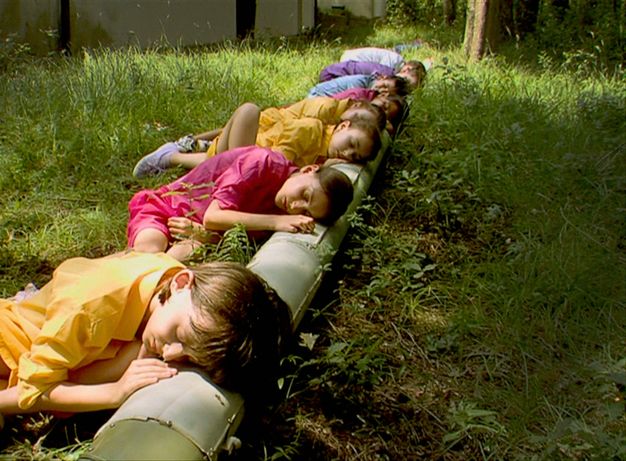 Video still. Eight children with pale or light brown skin lie with their eyes closed in a grassy field, their heads on a pale green pipe, which is used like a long pillow. The children wear clothing in vivid yellow, pink, blue, or purple.