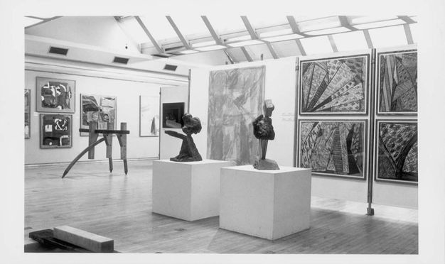 Installation photograph. This black and white photograph of two sculptures, possibly abstracted busts, on pedestals. Farther from us, another sculpture sits on pointed feet directly on the floor in a gallery hung with paintings or other media.